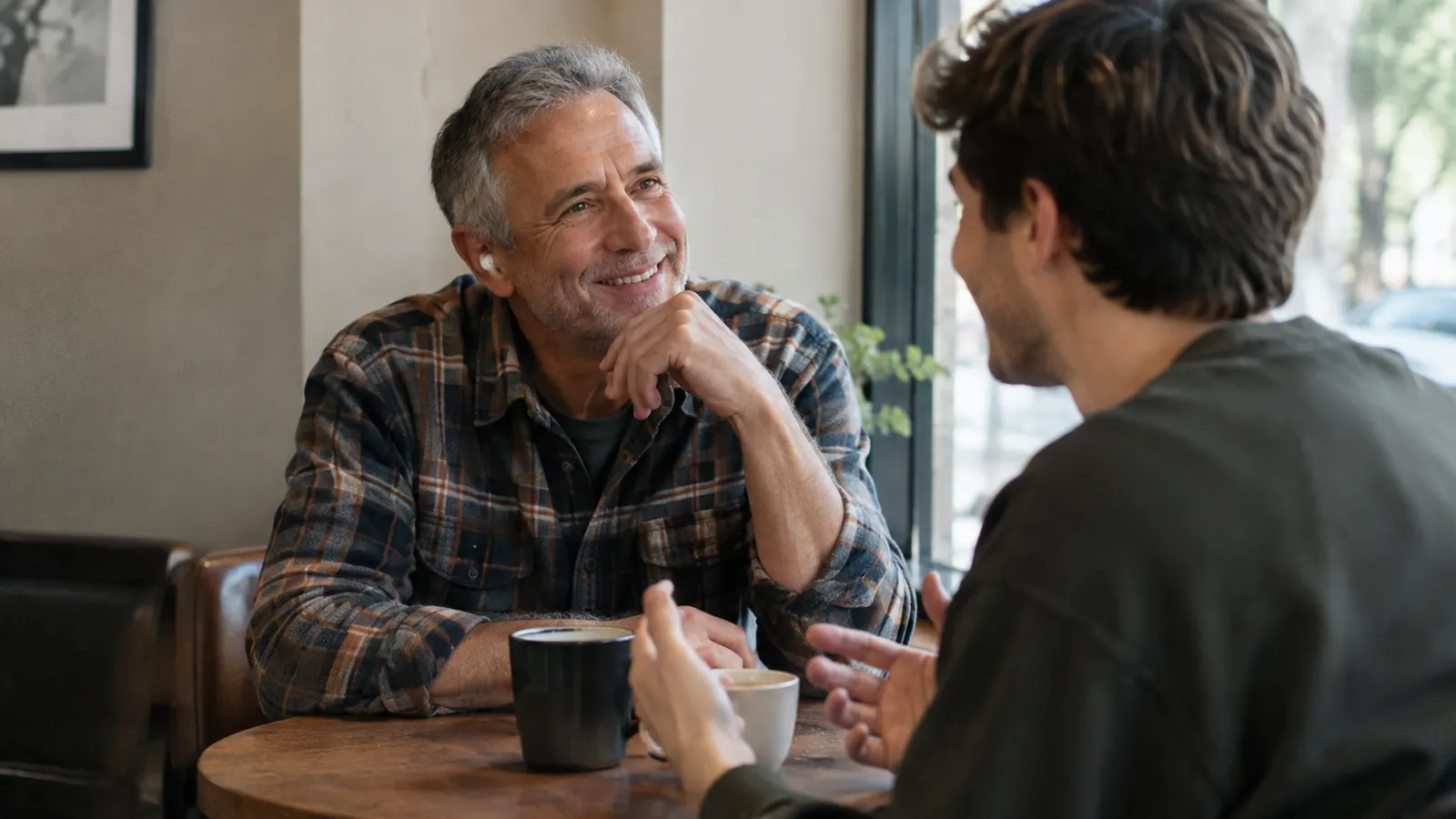 an older man wearing small earbuds smiling as he listens to his grandson in a quiet corner of a coffee shop